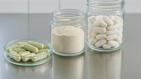 Close-up of supplement capsules, tablets, and powder in clear glass containers on a stainless steel surface.